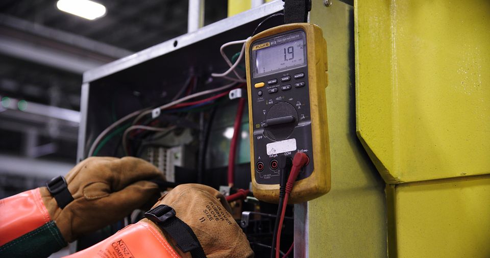 An electrician's gloved hands testing a panel with a multimeter.