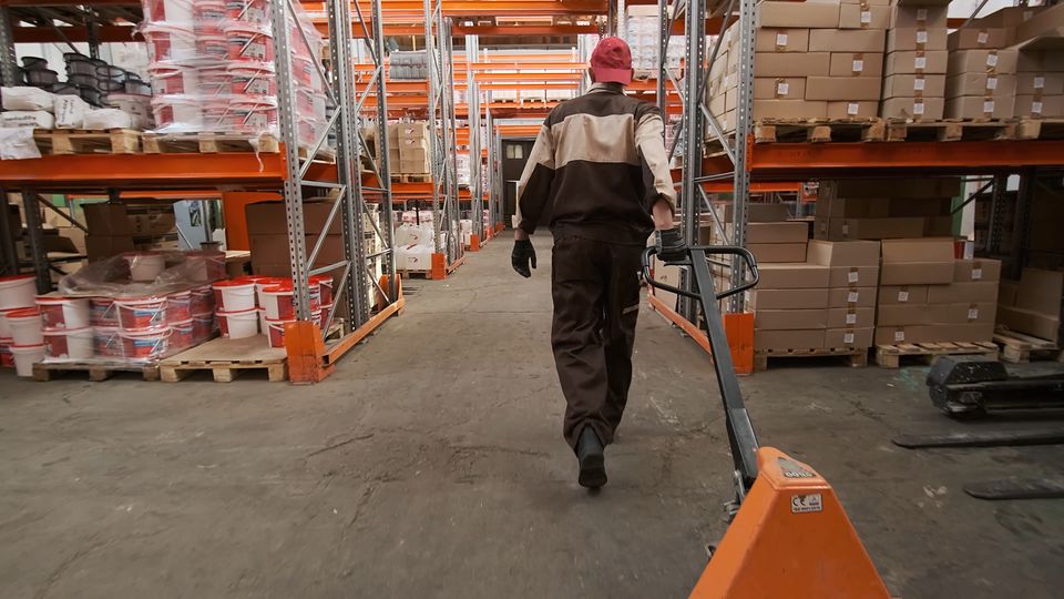 A warehouse worker pulling a pallet jack between tall shelves.